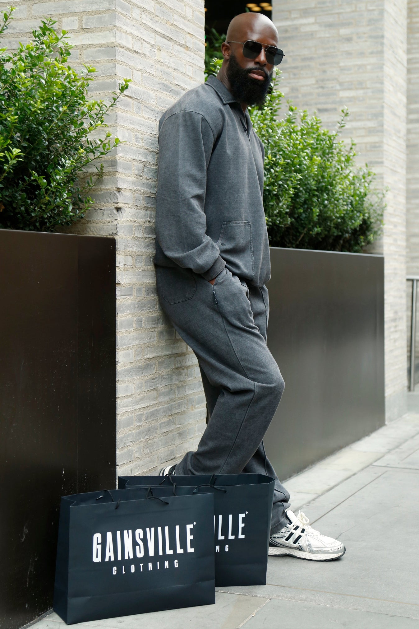 Man in charcoal gray Gainsville tracksuit leaning against a wall with 'Gainsville Outfitters' bags on the ground.